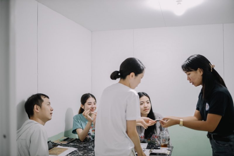 e-Room 的小教室裡，穿黑色 polo 衫的老師把東西遞給站著的學員，另外三位學員坐在桌旁，桌上有講義和飲料