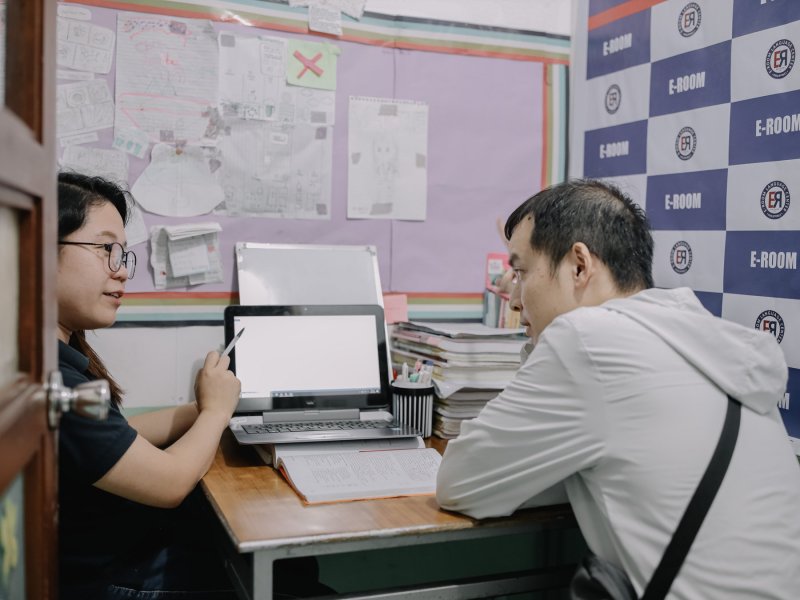 e-Room 一對一英文課,戴眼鏡的老師拿筆指著筆電講解,學生坐在對面聽,右側是 E-ROOM 校徽背板