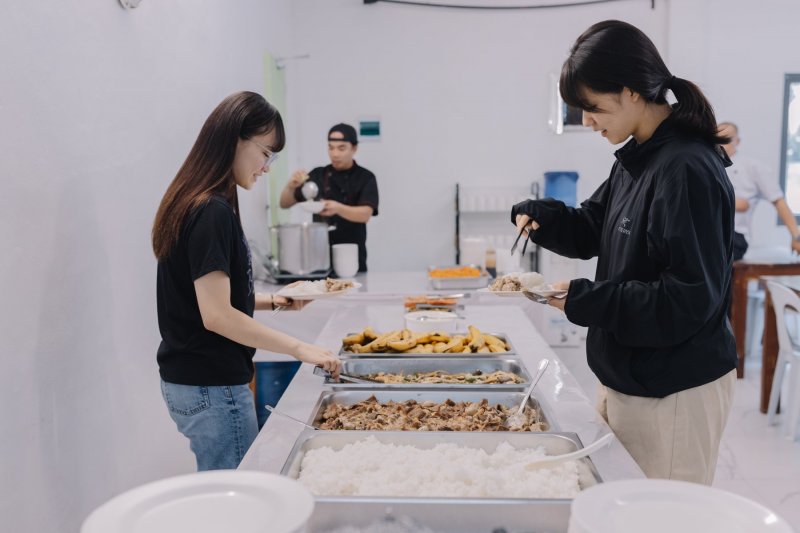 e-Room 學校餐廳的自助餐檯,兩位學生拿著盤子夾菜,檯上有白飯、炒肉等菜色,後方廚師正在盛湯