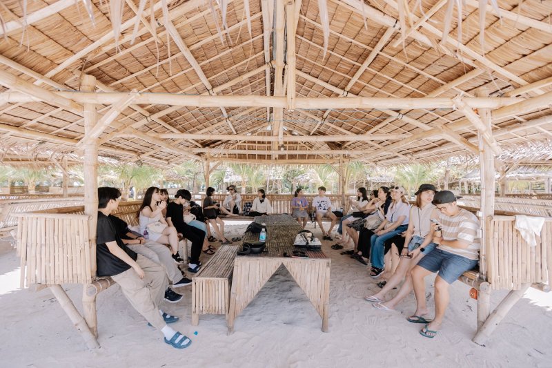 一群學生在拉卡旺島(Lakawon)海灘的竹編茅草涼亭裡休息聊天,地上是白沙,涼亭外有椰子樹