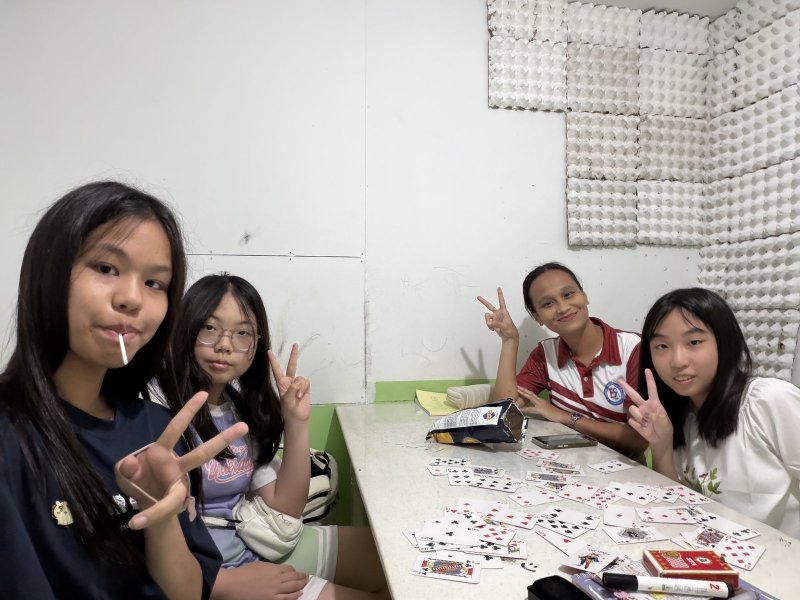 e-Room 教室裡，三個女孩和穿 e-Room polo 衫的女士圍著桌子玩撲克牌